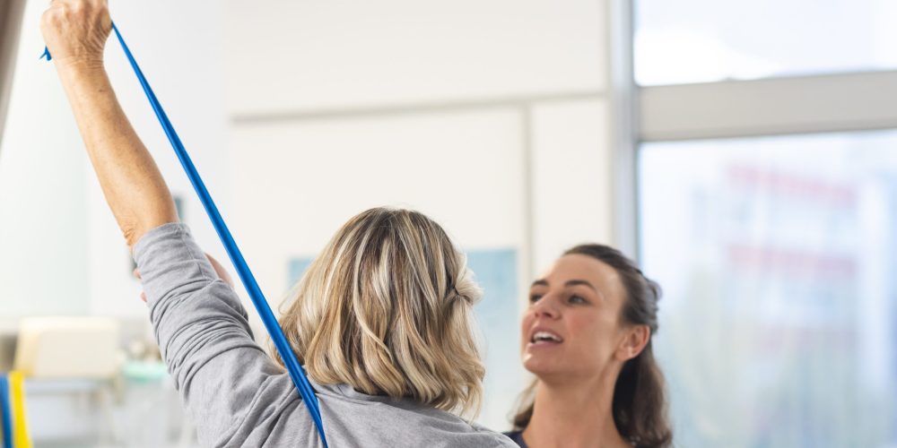 Caucasian female physiotherapist and senior woman using exercise band stretching arms